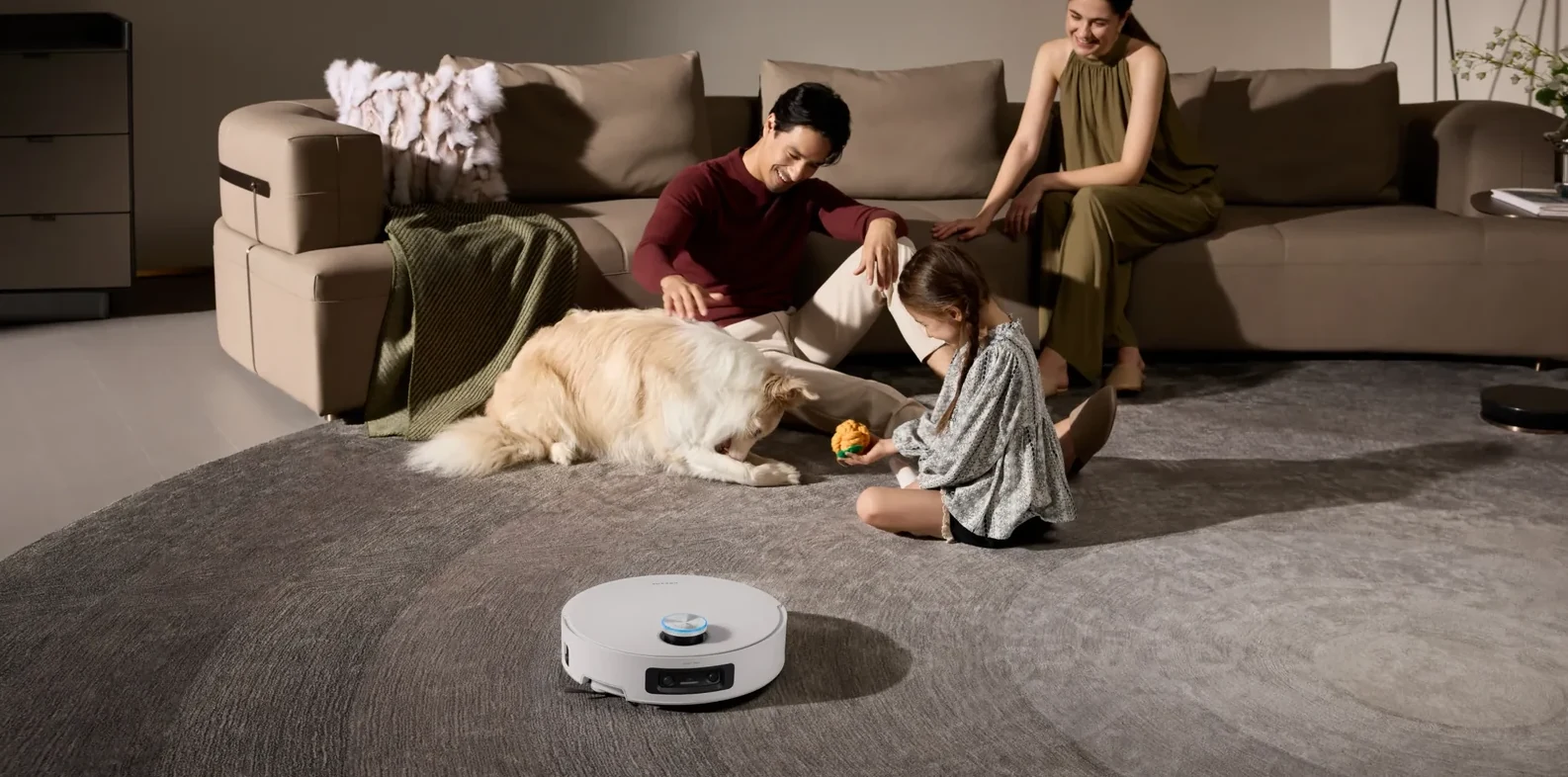 Šeima sėdi ant kilimo, robotas valo palei kraštą