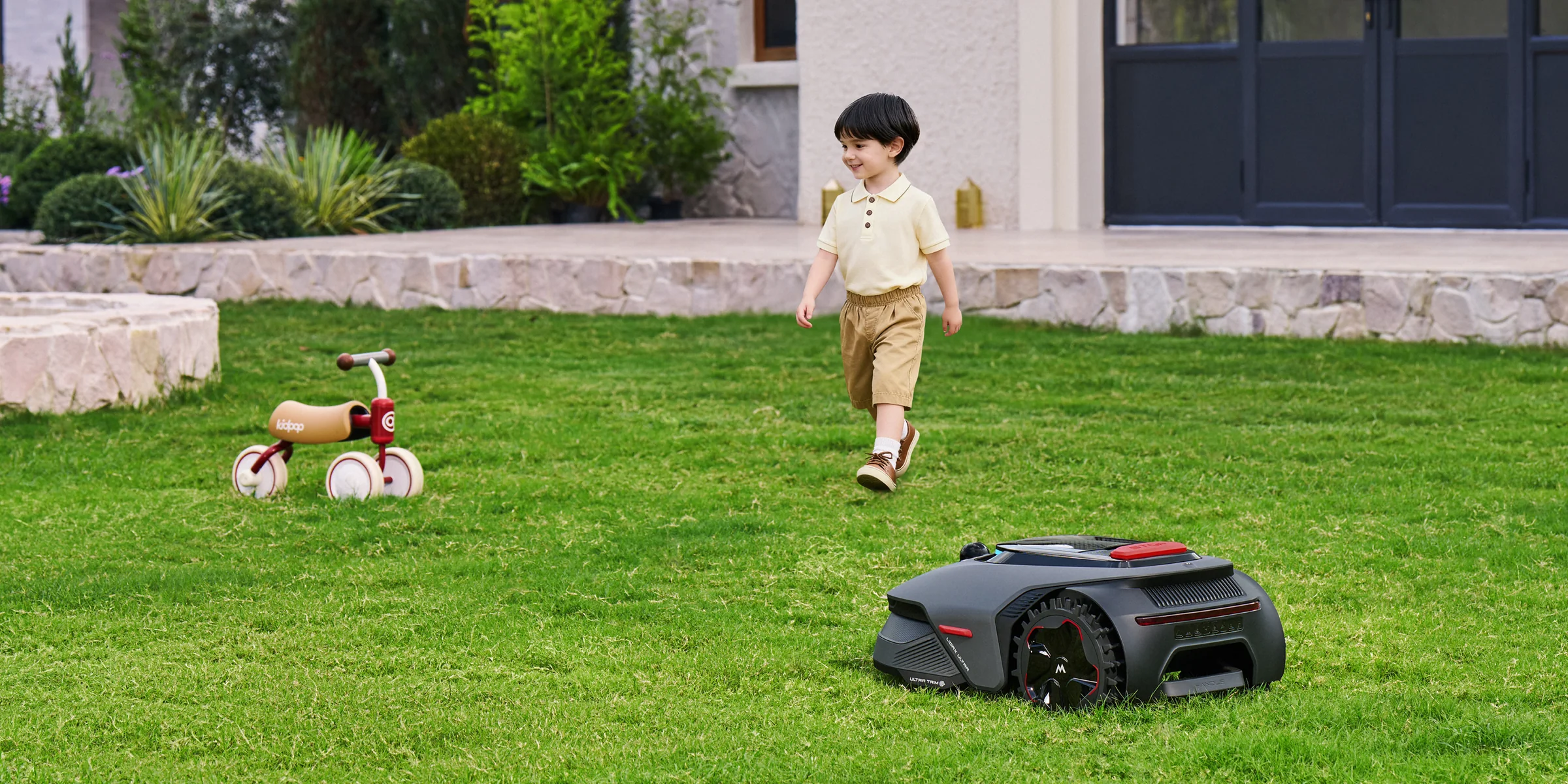 Vaikas žaidžia vejoje, šalia dirba vejos robotas