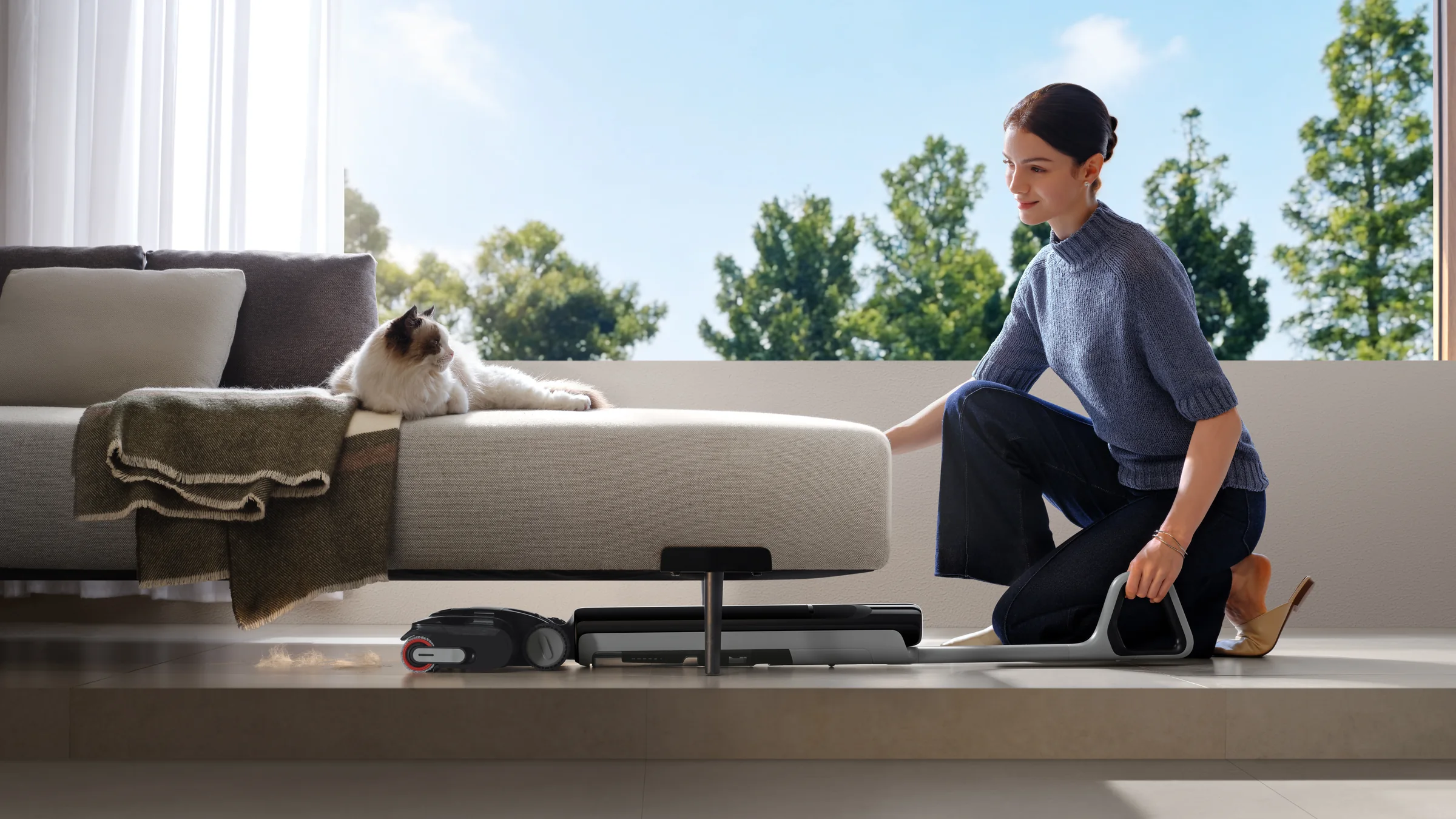 Moteris paguldytu DREAME G12 Dual siurbliu plauna grindis po žema sofa, ant sofos guli katė